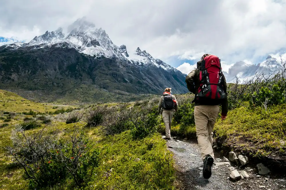 Two hikers with backpacks walking a trail through green alpine meadows toward snowy peaks