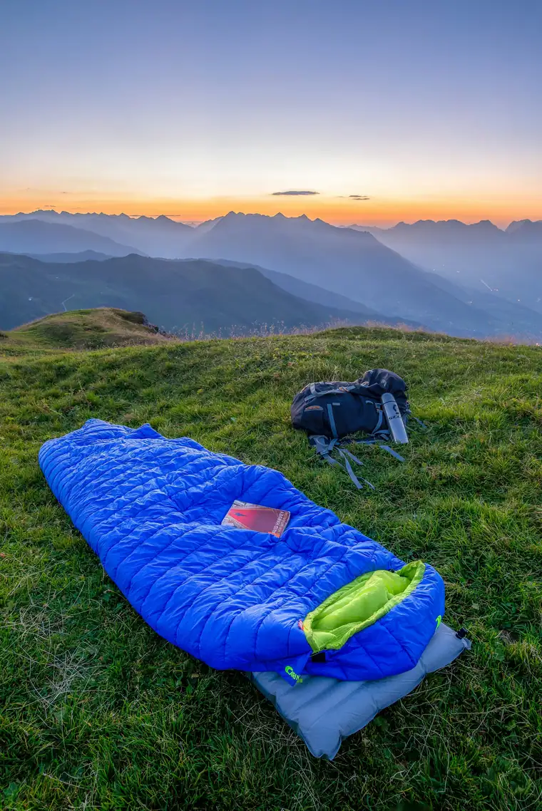 Blue sleeping bag and a small backpack laid out on a grassy mountain slope