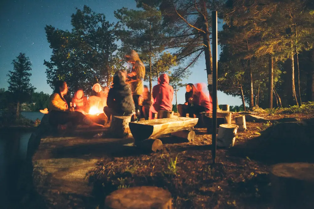 Small group of campers relaxing around a campfire among tall pine trees at dusk